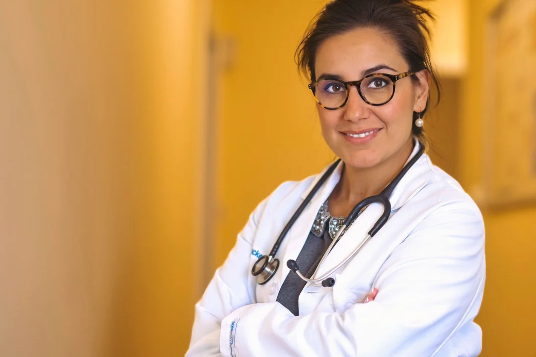 Reyhan Berkel, MD, in a white coat with a stethoscope around her neck and her arms folded, standing in the marigold corridor of her office.